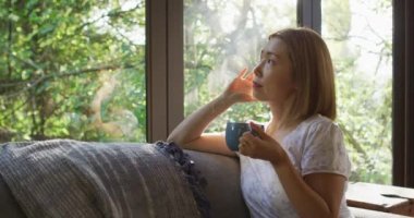 Pencereden bakan Asyalı bir kadın ve elinde bir kupa kahveyle kanepede yatıyor. Karantina süresince evde tecrit altında..