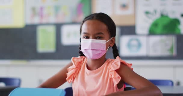 身穿面罩的混血女学生的画像 坐在教室里看着相机 Coronavirus Covid 19大流行病期间小学儿童 — 图库视频影像