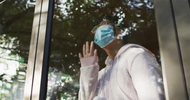顔のマスクをしたアジア系の少女が窓から眺めている 隔離中に隔離された家で — ストック動画