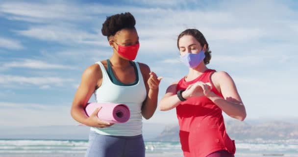 ビーチでヨガマットを持ってフェイスマスクを身に着けている2人の多様な女性の友人 健康的なアクティブライフスタイル屋外フィットネス健康 — ストック動画