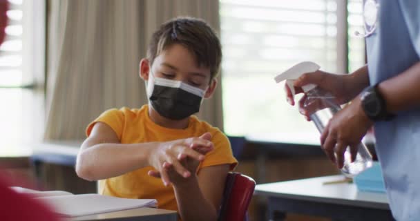 教室で手を消毒し 顔のマスクを身に着けている混合レースクールボーイの肖像画 コロナウイルス感染症の小学生の子供たちは — ストック動画