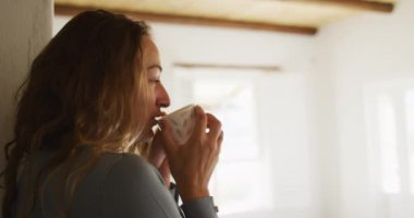 Güneşli yazlık mutfakta pencerenin yanında durup kahve içen düşünceli beyaz kadın. Kırsal kesimlerin dışında basit bir evde yaşamak.