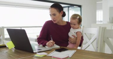 Beyaz bir anne bebeğini kucağında tutuyor ve evde çalışırken not alıyor. annelik, sevgi ve çocuk bakımı kavramı