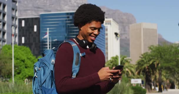 スマートフォンを使ってアフリカ系アメリカ人の笑顔 街の通りでヘッドフォンやバックパックを着用 外にも街中にもデジタル ノマドが — ストック動画