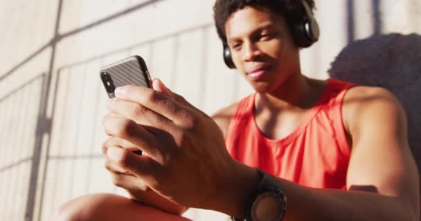 市内の幸せなアフリカ系アメリカ人男性は ワイヤレスヘッドフォンでスマートフォンを使用して 壁に座っています フィットネスとアクティブな都会のアウトドアライフスタイル — ストック動画