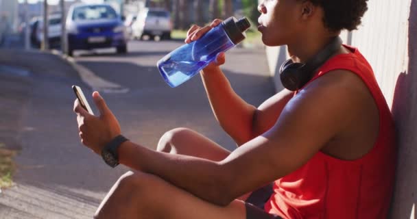 アフリカ系アメリカ人の男性に合う都市で休憩を取り スマートフォンを使用して 飲料水を座って運動 フィットネスとアクティブな都会のアウトドアライフスタイル — ストック動画