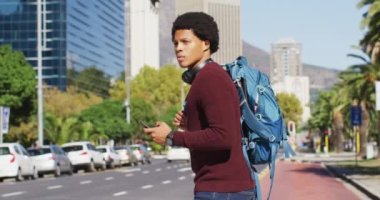 Şehirde Afro-Amerikan bir adam, akıllı telefon kullanıyor, kulaklık takıyor ve sırt çantasıyla karşıdan karşıya geçiyor. Dijital göçebe şehirde geziyor, dolaşıyor..