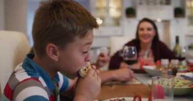 Aile yemeği sırasında masada yemek yiyen gülümseyen beyaz bir oğul, arka planda ebeveynler. Aile evde eğleniyor..