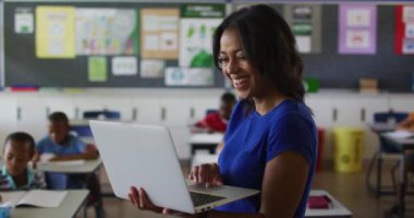 Sınıfta dizüstü bilgisayarlı çocuklarla duran mutlu melez kadın öğretmenin portresi. İlkokuldaki çocuklar.