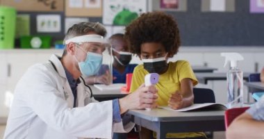 Çeşitli erkek öğretmenler okul çocuğuna vücut ısısını nasıl ölçeceğini gösteriyor, hepsi yüz maskesi takıyor. Coronavirus covid 19 salgını sırasında ilkokuldaki çocuklar.