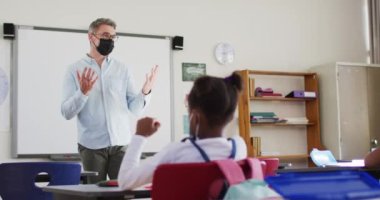 Yüz maskesi takan beyaz erkek öğretmen okulda öğrencilere ders veriyor. Covid 19 salgını sırasında okulda hijyen ve sosyal mesafeler.