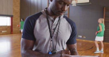 Arka planda çeşitli bayan basketbol takımları olan Afrika kökenli Amerikalı erkek koçun portresi. Basketbol, kapalı sahada spor eğitimi..