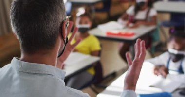 Sınıfta çeşitli erkek öğretmenler ve çocuklar ders sırasında el kaldırıyor, hepsi yüz maskesi takıyor. Coronavirus covid 19 salgını sırasında ilkokuldaki çocuklar.
