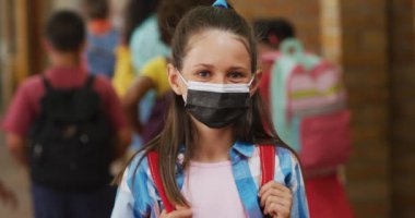 Kafkas öğrenci portresi yüz maskesi takıyor, koridorda dikilip kameraya bakıyor. Coronavirus covid 19 salgını sırasında ilkokuldaki çocuklar.