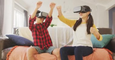 Mutlu Asyalı erkek ve kız kardeşler evlerinde, oturma odasındaki kanepede VR kulaklıklar takarak oturuyorlar. Karantina süresince evde tecrit altında..