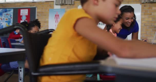 教室でノートを作る 背景に教師に座って 車いすの中で混合レースの学校の少年 小学校の子どもたちは — ストック動画