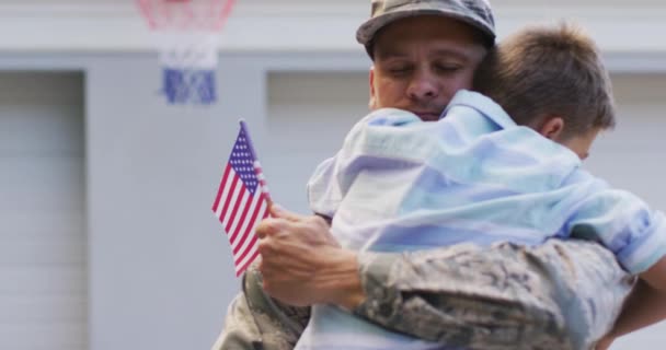 家の外の庭に旗を掲げて笑顔の息子を抱きしめる幸せな白人男性兵士 兵士は家族の元に戻り — ストック動画