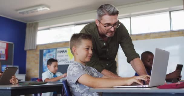 ラップトップを使って教室に座っている男の子を助ける多様な男性教師 小学校の子どもたちは — ストック動画