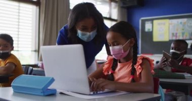 Çeşitli bayan öğretmenler öğrencilere yardım ediyor, hepsi de yüz maskesi takıyor. Coronavirus covid 19 salgını sırasında ilkokuldaki çocuklar.