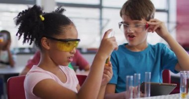 Kimya dersi sırasında çeşitli ırk öğrencileri koruyucu gözlük takıyor ve ellerinde tüp var. İlkokuldaki çocuklar.