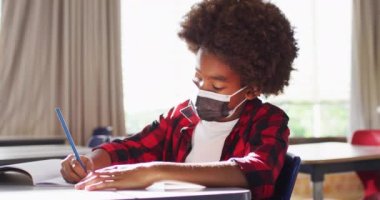 Sınıfta kameraya bakarken yüz maskesi takan Afro-Amerikan okul çocuğunun portresi. Coronavirus covid 19 salgını sırasında ilkokuldaki çocuklar.