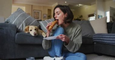 Gülümseyen beyaz kadın kruvasan yiyor ve yanındaki koltukta köpeğini okşuyor. evcil hayvan arkadaşlığı, ev hayatı ve ev konseptinden çalışma.