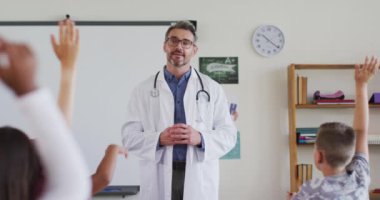 Setoskop takan beyaz bir erkek doktor sınıfta dikilip sorular soruyor. İlkokuldaki çocuklar.