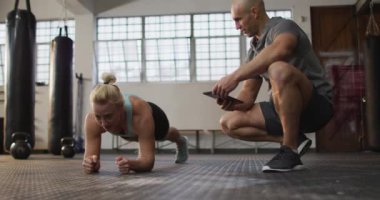 Spor salonunda elinde dijital tablet tutan erkek antrenörle tahta egzersizi yapan beyaz bir kadın. Spor, antrenman ve spor konsepti