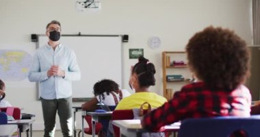 Sınıfta çeşitli erkek öğretmenler ve çocuklar ders sırasında el kaldırıyor, hepsi yüz maskesi takıyor. Coronavirus covid 19 salgını sırasında ilkokuldaki çocuklar.