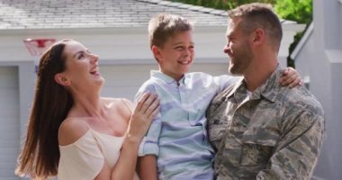 Mutlu beyaz erkek asker, karısının bahçesinde gülümseyen oğlunu kucaklıyor. Asker evine, ailesine dönüyor..