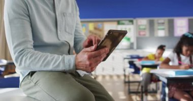 Sınıfta, kameraya bakan tablet kullanan çocuklarla birlikte mutlu beyaz erkek öğretmenin portresi. İlkokuldaki çocuklar.