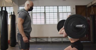 Yüz maskesi takan beyaz bir kadın spor salonunda erkek antrenörle halterle çalışıyor. Coronavirus covid-19 pandemik konsepti sırasında spor, eğitim ve spor