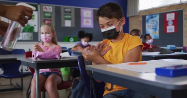 教室で手を消毒顔マスクを身に着けている混合レースの学童は バックグラウンドで同僚 コロナウイルス感染症の小学生の子供たちは — ストック動画