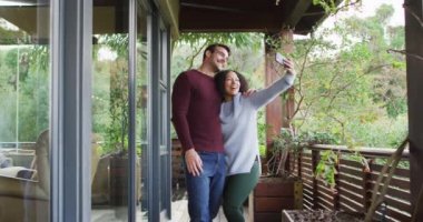 Karışık ırk çifti gülümserken yazlık evin balkonunda akıllı telefondan selfie çekiyor. çift balayı ve tatil konsepti