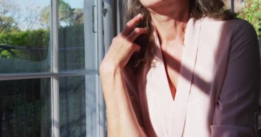 Düşünceli son sınıf beyaz kadın güneşli oturma odasında oturmuş pencereden dışarı bakıyor. emeklilik yaşam tarzı evde.