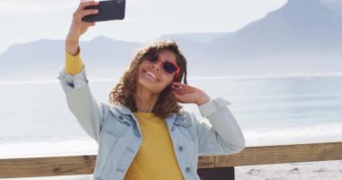 Mutlu melez kadın selfie çekiyor ve deniz kenarındaki güneşli gezinti alanında kolları havada gülümsüyor. Boş zaman, açık hava ve doğaya yakın..