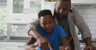 Mutlu Afro-Amerikan babası, oğlunun üzerine eğilip mutfak masasına oturuyor ve ödevlerine yardım ediyor. Aile evde birlikte vakit geçiriyor..
