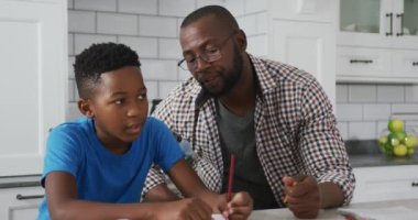 Afrika kökenli Amerikalı baba mutfak masasında oturup oğluna ödevlerinde yardım ediyor. Aile evde birlikte vakit geçiriyor..