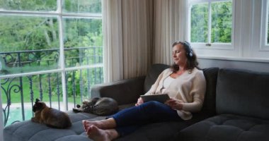 Video konuşması, konuşması ve kediyi okşaması için kulaklık ve tablet kullanan Kafkasyalı mutlu bir kadın. emeklilik yaşam tarzı evde iletişim teknolojisi ile.