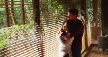 Happy diverse couple embracing and smiling in living room, looking out of window in the countryside. spending free time together at home.