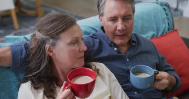 リビングルームのコーヒーを飲みながら話しているソファの上に座っているシニア白人のカップル 家庭での定年退職生活 — ストック動画