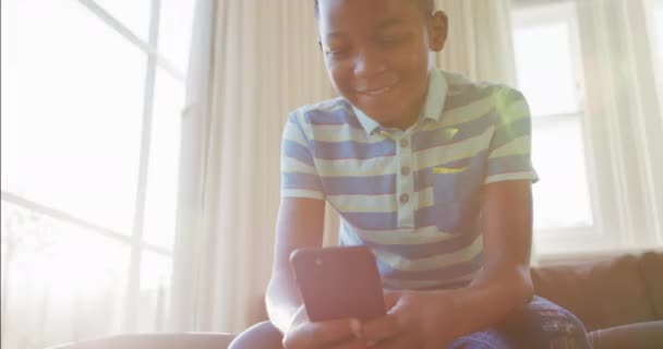 Χαρούμενο Αφροαμερικανό Αγόρι Που Κάθεται Στον Καναπέ Χρησιμοποιώντας Smartphone Και — Αρχείο Βίντεο