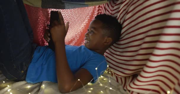 幸せなアフリカ系アメリカ人の少年は タブレットを使用して 毛布のキャンプに横たわって笑顔 家族で家で過ごす時間は — ストック動画