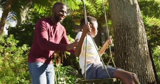 アフリカ系アメリカ人の父親は 明るい晴れた日に庭でスイングに彼の息子を押しています 父親と愛の概念 — ストック動画