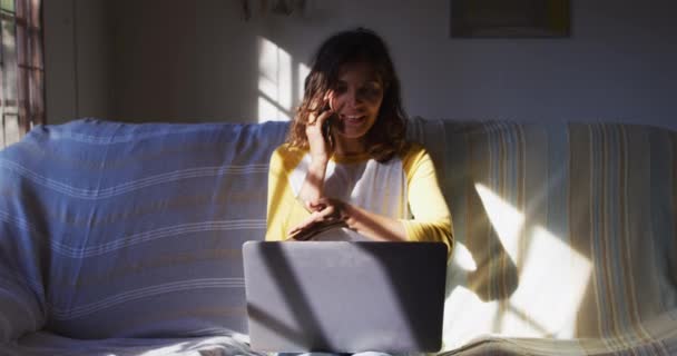 快乐的混血女子坐在沙发上 在阳光充足的客厅里用笔记本电脑用智能手机聊天 隔离检疫期间在家中单独工作 — 图库视频影像