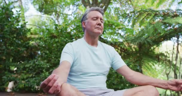 Entspannter älterer kaukasischer Mann praktiziert Yoga sitzend in Lotusposition und meditiert im Garten. Gesunder Lebensstil im Ruhestand.