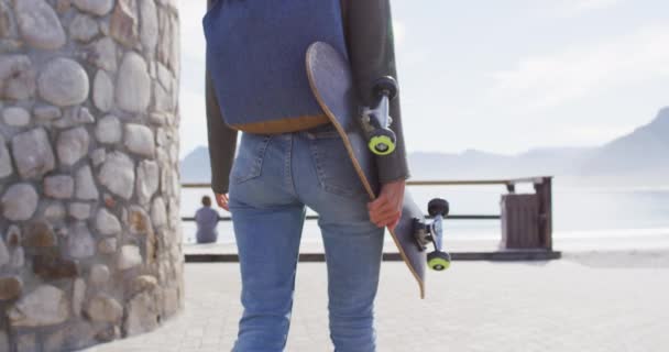 海沿いの遊歩道を歩くスケートボードを保持する混合レース女性の後部ビューミッドセクション 健康的な生活を送っています — ストック動画