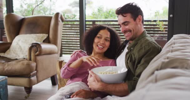 休暇の家で一緒に映画を見ながら 混合レースのカップルはポップコーンを食べて笑顔 新婚旅行と休暇のコンセプトを — ストック動画