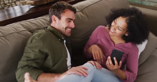 休暇の家でソファの上で一緒にスマートフォンを見ている間に混合レースのカップル笑顔 新婚旅行と休暇のコンセプトを — ストック動画