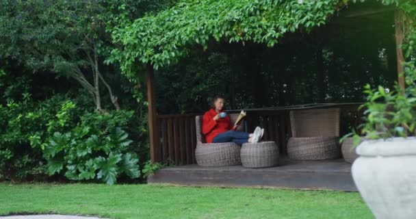 シニア白人女性は庭でリラックスし 足で座ってコーヒーを飲み 本を読む 家庭での退職生活だけで — ストック動画